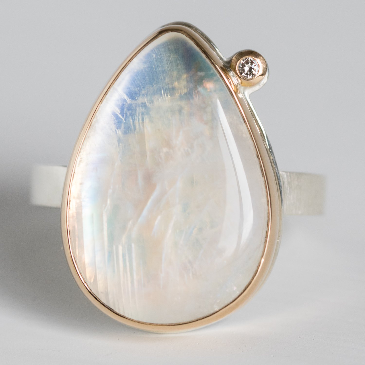 Jamie Joseph: 14K gold and sterling silver ring with vertical teardrop smooth white rainbow moonstone and .03ct satellite diamond on X band Size 7.5